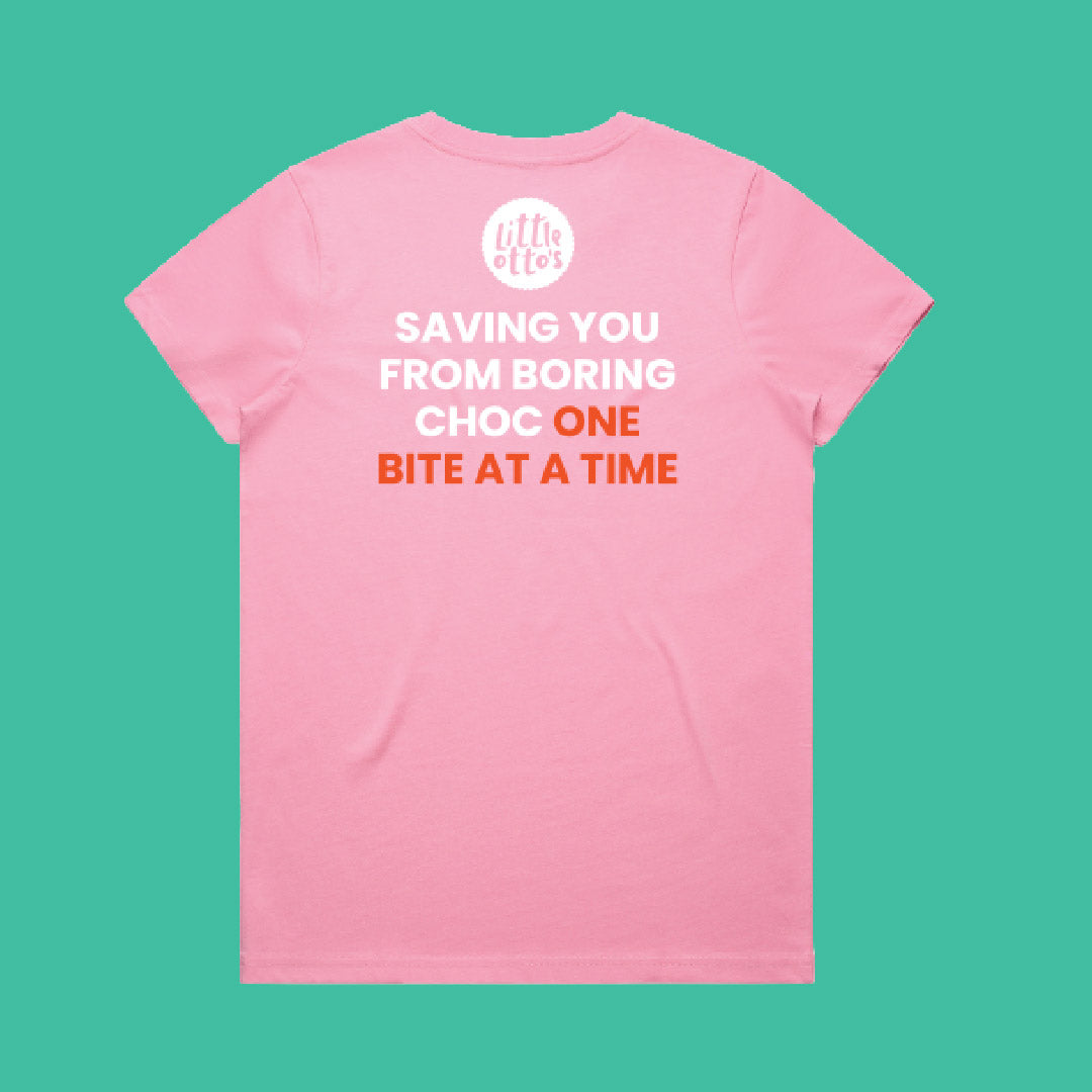 A pink short-sleeve t-shirt with a white and orange graphic print on the back, displayed flat against a solid teal background. The graphic features a white circular logo at the top and the text 'SAVING YOU FROM BORING CHOC ONE BITE AT A TIME' in white and orange lettering.