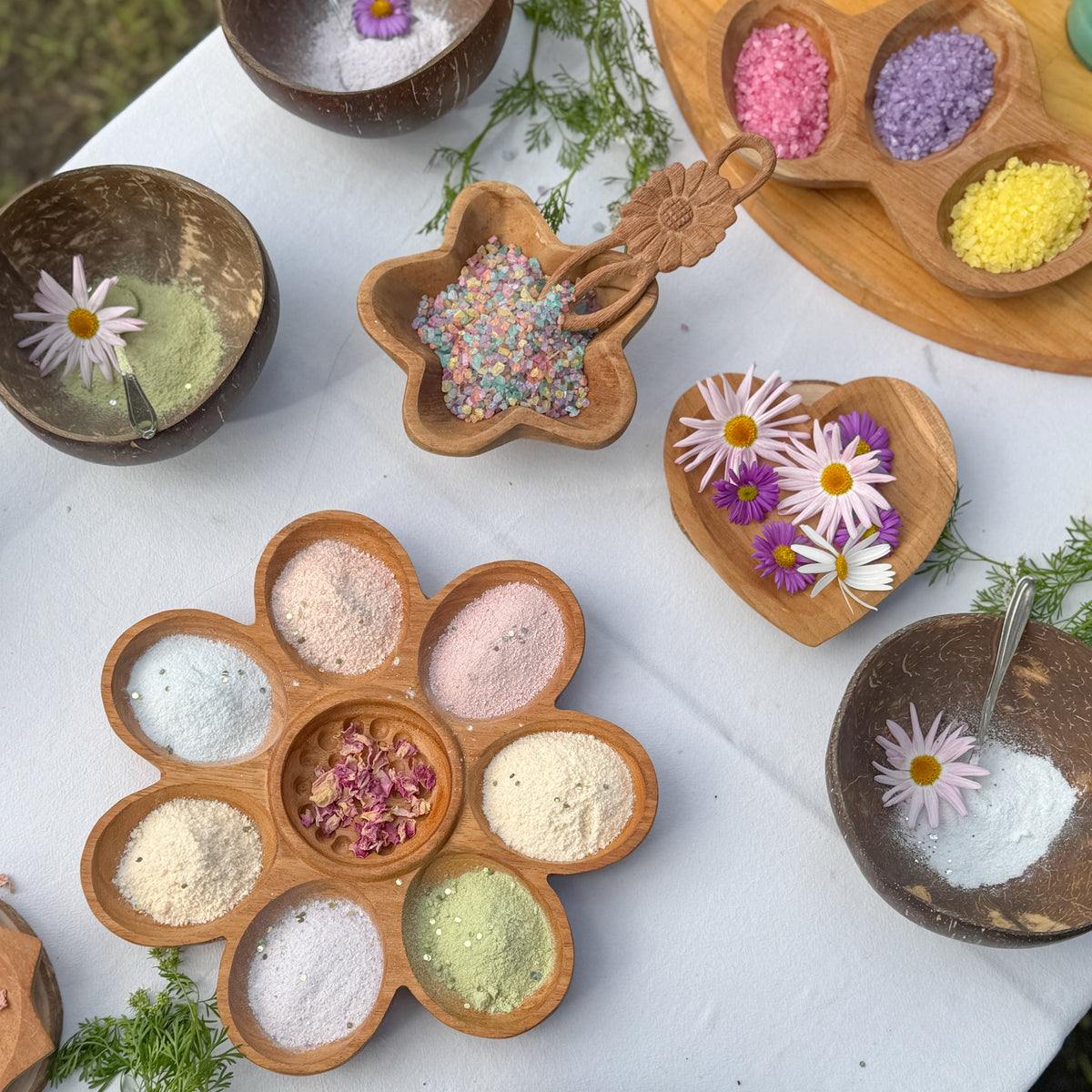 An overhead styled flat-lay of multiple small bowls and wooden trays filled with colorful powders, crystals, and flower petals on a white cloth. No person or animal is present, and the scene is arranged as a product display rather than a real-use environment, so Product/Packaging fits better than Lifestyle.